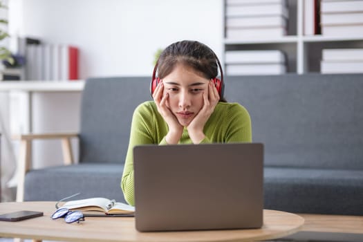 Asian woman feeling stressed while working on laptop with headphones in modern office, concept of stress.