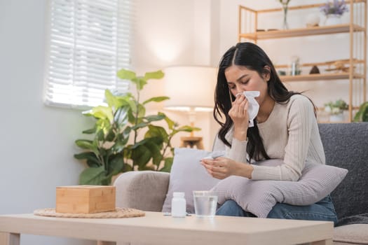 A young woman sitting on a couch in a bright living room, blowing her nose with a tissue. Plants and shelves are visible in the background.