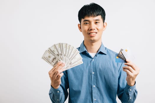 A confident Asian man smiles while holding dollars and a credit card for payment in his hand. Studio shot isolated on white background, showcasing financial success.