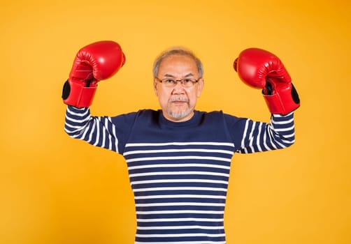 Portrait Asian old man wearing glasses wear two red boxing gloves studio shot isolated yellow background, smiling happy elderly man gray haired healthy fighter lifestyle concept