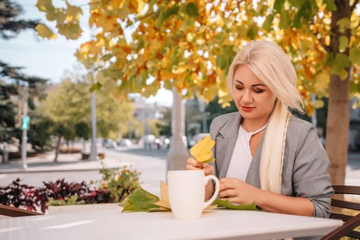 A blonde woman sits at a table with a cup of coffee and a leaf on it. The scene is set in a city with a tree in the background