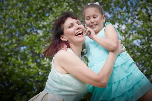 Happy mother and daughter enjoying rest, playing and fun on nature on a green lawn and with blooming apple tree in the background. Woman and girl resting outdoors in summer and spring day