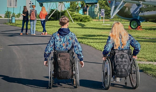 Two individuals, a man and a woman, both in wheelchairs, are exploring an outdoor museum featuring retired military helicopters on a sunny day, showcasing accessibility and inclusion.