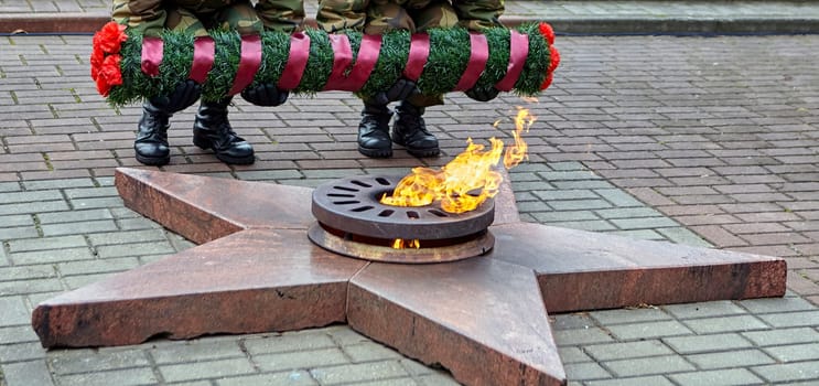 Two soldiers in uniform standing solemnly behind a wreath with red and black ribbons, in front of an eternal flame burning in a star-shaped monument at a military ceremony.