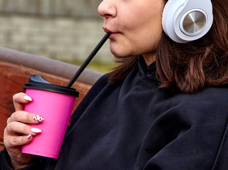 A young woman with long brown hair and white headphones is drinking from a pink cup with a straw. She is wearing a black sweatshirt and has her nails painted pink with white polka dots.