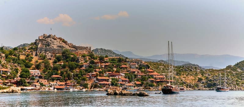 Wooden sailing boat in calm sea with green mountains. Two masts, white sails. Clear blue-green water, blue sky, white clouds. Marina with more boats. Great for travel brochure.