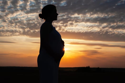 The silhouette of a pregnant woman at sunset with her hands on her belly. The sky is ablaze with color, and the clouds are forming a beautiful pattern. A truly stunning and peaceful scene.