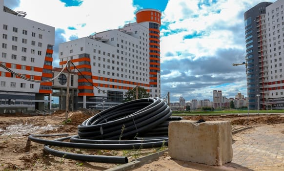 A pile of black pipes sits in front of two unfinished apartment buildings, with a concrete block in the foreground. Construction cranes are visible in the background, while a cityscape of other buildings stretches out under a partly cloudy sky.