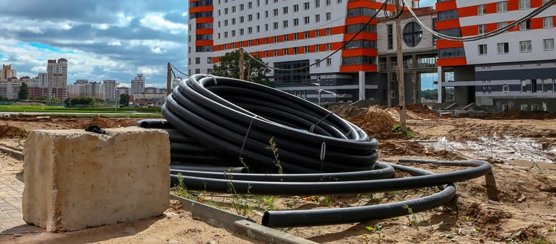 A large coil of black pipes sits on a dirt construction site, resting against a concrete block. The pipes appear to be part of a new infrastructure project, with a newly constructed apartment building in the background.