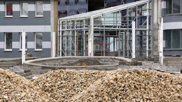 A modern building under construction with a large glass facade. The structure is incomplete with visible scaffolding and piles of construction aggregate in the foreground.
