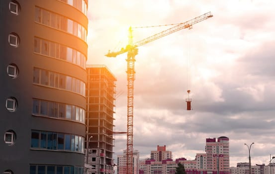 A construction crane stands tall against a backdrop of a city skyline, silhouetted by the setting sun. Buildings are under construction, showing the progress of urban development.