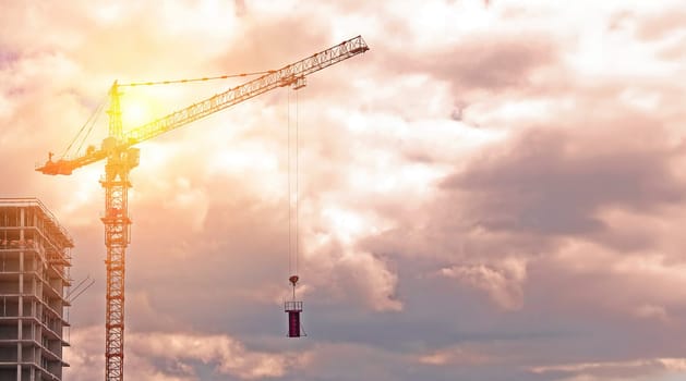 A construction crane stands tall against a backdrop of soft pink and white clouds, with a building under construction visible in the foreground.