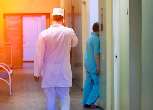 A doctor in a white coat walks down a hospital hallway, with a patient waiting in the background. The doctor is moving quickly and decisively.