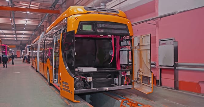 A bright orange electric bus is being assembled in a factory, with its doors open and various wires and components visible. The bus is positioned on a platform with other buses behind it, creating a sense of production and progress.