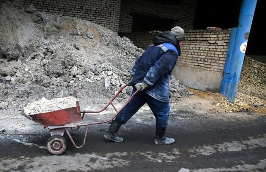 A man in a blue jacket and work boots pushes a wheelbarrow filled with gravel along a dirt path. The path leads to a brick wall and a pile of gravel.