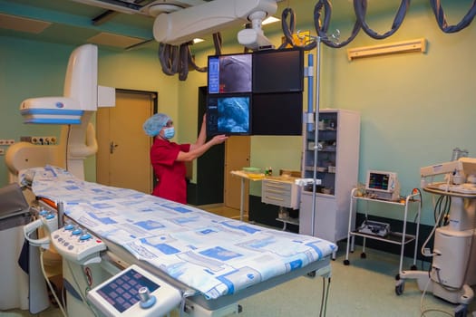 A medical professional in a hospital operating room monitors a patient during a diagnostic imaging procedure. The room is equipped with state-of-the-art equipment, including a large display screen displaying images from the procedure.