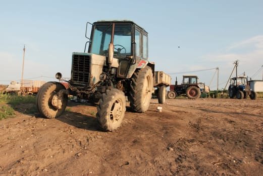 A weathered tractor sits in a rural field under a clear blue sky. The tractor is covered in rust, and its tires are worn down. The surrounding field is covered in dirt and grass, and there are other tractors in the distance.