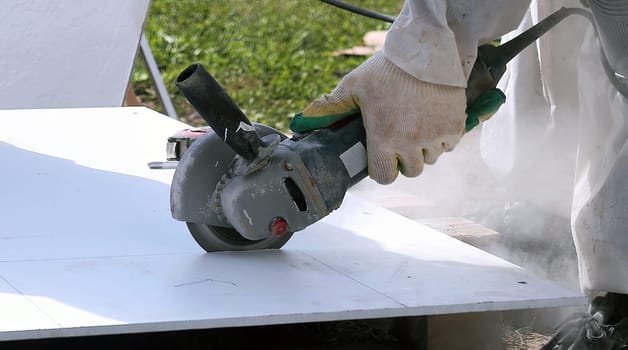 A person wearing gloves uses an angle grinder to cut through a white material outdoors. Dust and sparks fly up as the grinder spins.