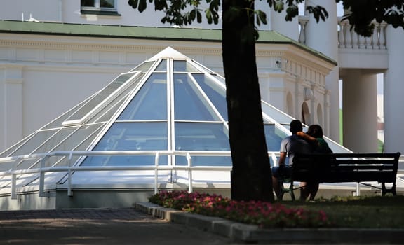 A couple sits on a bench, embracing under the shade of a large tree, with a glass pyramid structure in the background. The sun casts a warm glow on the scene, creating a peaceful and romantic ambiance.