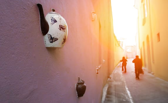 A white teapot with butterfly designs is attached to a pink wall in a narrow alleyway. Two figures walk away from the camera in the background, as the sun shines brightly at the end of the street.