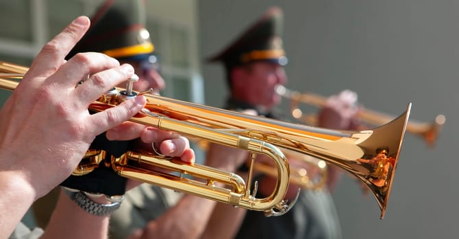 A close-up view of a trumpeters hands, playing their instrument during a daytime parade. The sunlight glints off the brass instrument and illuminates the scene.