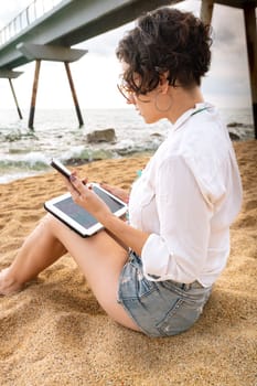 Caucasian woman on the beach with digital tablet and smartphone in hand typing smiling with sunglasses