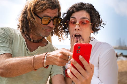 Middle-aged man and woman sitting on the beach with surprised face, browsing smartphone apps. Concept:Holidays y Technology