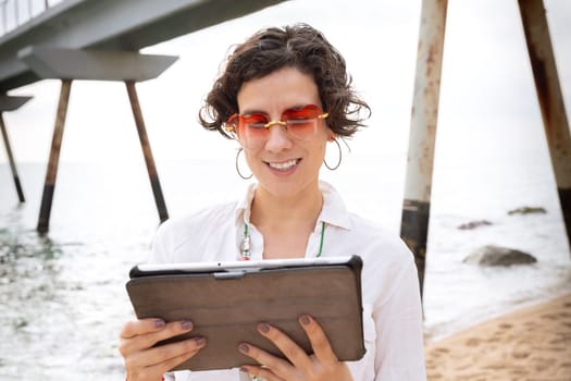 Woman on the beach with digital tablet in hand typing smiling with sunglasses