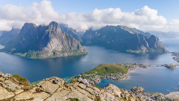 Reine in Norway, with a couple sitting on a cliff overlooking the stunning landscape of mountains, fjords, and islands. a couple on top of a mountain at Reinebringen, Lofoten, Norway