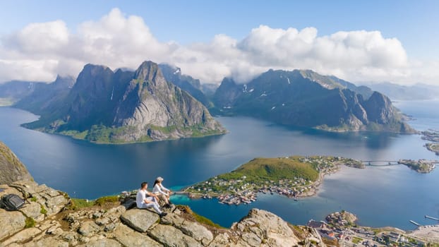 A scenic view from a mountaintop in Norway, showcasing a stunning fjord surrounded by majestic mountains. Two figures enjoy the panorama. a couple of men and women at Reinebringen, Lofoten, Norway