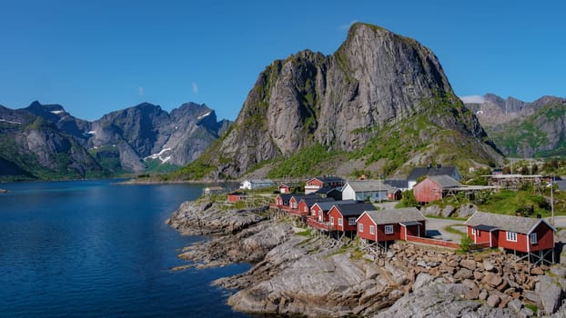 A scenic view of red cabins situated on the rocky shores of a fjord in Norway. Surrounded by towering mountains, Hamnoy fishing village on Lofoten Islands, Norway with red rorbu houses