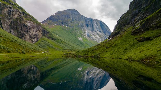 A serene lake nestled between towering mountains in Norway, reflecting the surrounding landscape. The sky is cloudy, adding to the atmosphere.