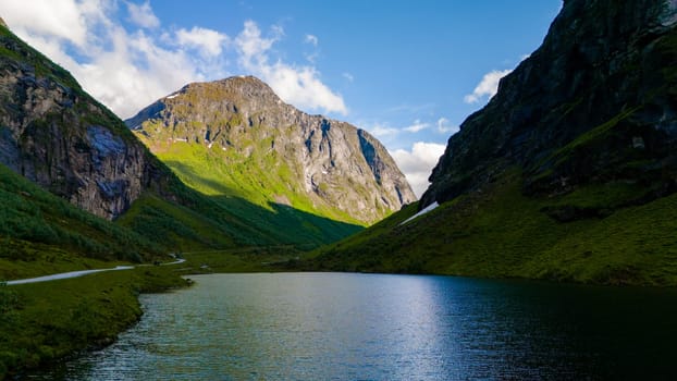 A tranquil lake surrounded by verdant hills and towering mountains in Norway. The calm waters reflect the blue sky and white clouds.