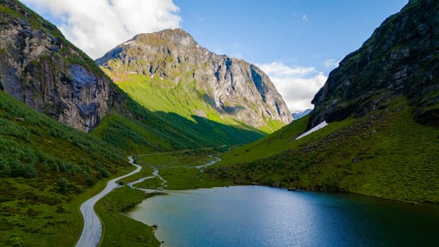 An aerial view of a winding road through a lush green valley in Norway, with a serene lake at the bottom and towering mountains on either side.