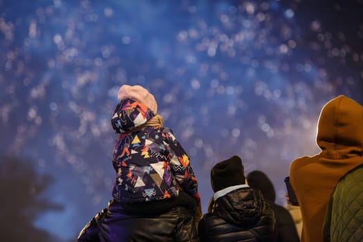 group of unrecognizable people with a child on shoulders in warm clothes are watching fireworks in a night sky, rear view