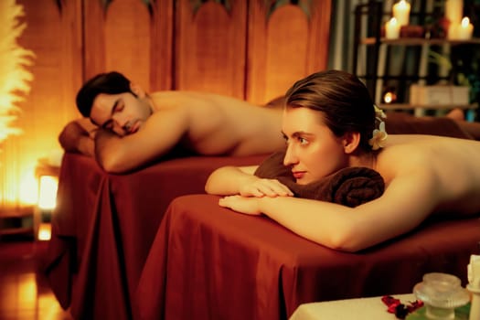 Caucasian couple customer enjoying relaxing anti-stress spa massage and pampering with beauty skin recreation leisure in warm candle lighting ambient salon spa at luxury resort or hotel. Quiescent