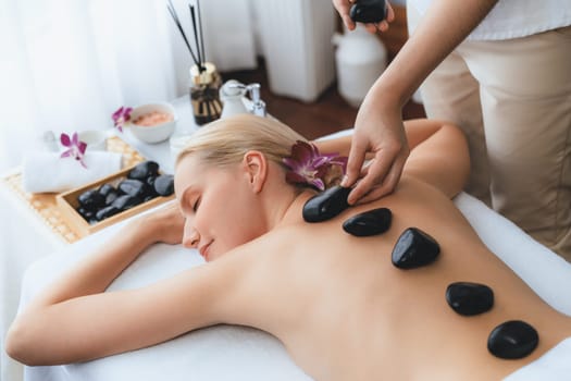 Hot stone massage at spa salon in luxury resort with day light serenity ambient, blissful woman customer enjoying spa basalt stone massage glide over body with soothing warmth. Quiescent