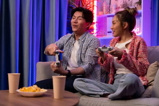 Sitting sofa of happy smiling couple playing video game control by joystick fronted snack and drinks competing challenge level with funny at modern neon blub light comfy living room at home. Infobahn.