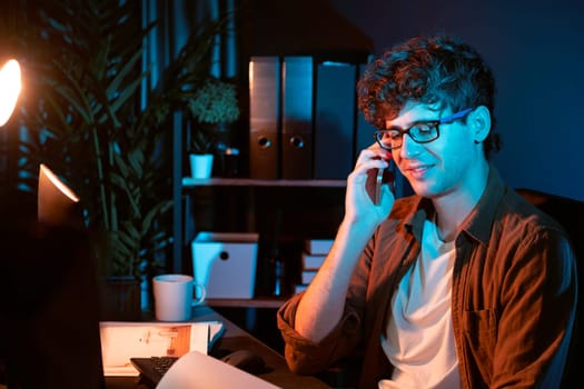 Young businessman calling on smartphone to customer or coworker, looking at computer screen of creative presentation surround work desk at neon modern office. Concept of new next project. Gusher.