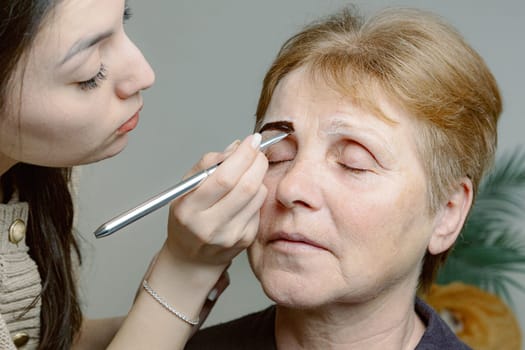One young Caucasian young girl cosmetologist carefully applies dark brown paint with a brush on the right eyebrow of an elderly woman with closed eyes, sitting on a white background in a home beauty salon, top side view very close up.