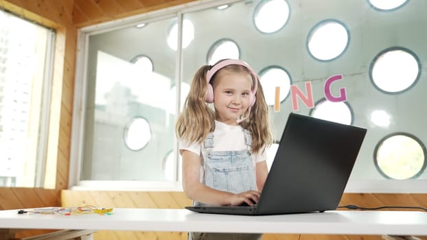 Little young cute girl using laptop to working or playing games. Student working on computer while looking and smiling at camera. Caucasian child attend online class by using headphone. Erudition.