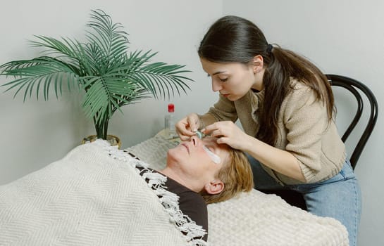 One Caucasian young beautiful brunette girl cosmetologist sticking a blue silicone eyelash stand on the right eye of an elderly client who is lying on a cosmetology bed in a home beauty salon, close-up side view.