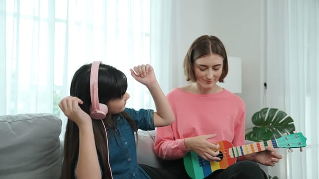 Asian cute girl listening to music from laptop by using headphone while lovely mom playing ukulele while teaching young daughter playing acoustic music. Cute child learning about instrument. Pedagogy.