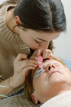 One Caucasian young beautiful brunette girl cosmetologist sticks combing the eyelashes of the right eye with a pink plastic brush to an elderly client who is lying on a cosmetology bed in a home beauty salon, bottom close-up view.