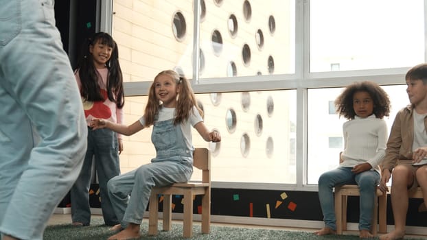 Diverse children playing musical chairs games while young smart beautiful teacher turn off music. Multicultural students running and sitting on chair in creative activity. Music lesson. Erudition.