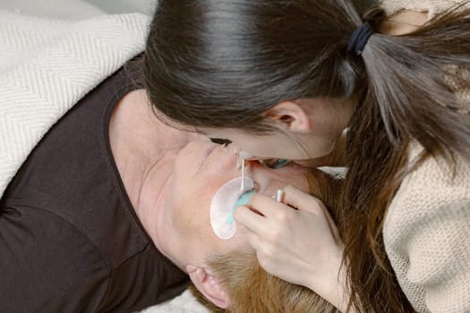 One Caucasian young beautiful brunette girl cosmetologist pulls out and straightens with a cotton stick the eyelashes of the right eye of an elderly female client who is lying on a cosmetology bed in a home beauty salon, top side view close-up.