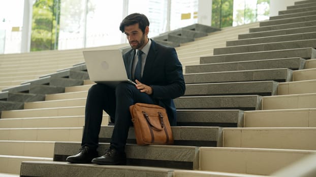 Skilled business man working by using laptop while sitting at stairs. Professional project manager typing and writing marketing plan. Handsome caucasian investor wearing suit with bag. Exultant.