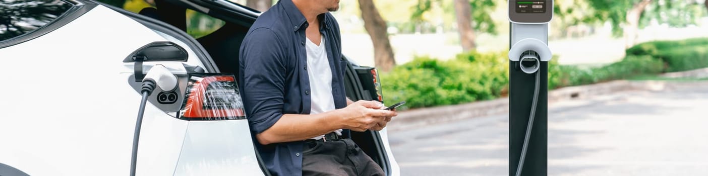 Man using smartphone online banking application to pay for electric car battery charging from EV charging station during vacation road trip at national park or summer forest. Panorama Exalt