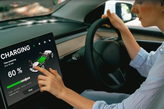 Young woman inside the vehicle, checking EV vehicle's application on battery recharging electricity status display on smart monitor screen in modern EV car on her road trip journey. Exalt