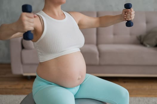 Pregnant woman doing exercises with dumbbells while sitting on a fitness ball at home. Close-up of a pregnant belly